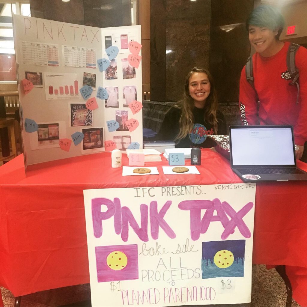 Two students smiling while tabling with a sign that reads: Pink Tax.