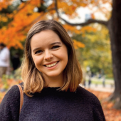 Headshot of Allegra Brochin, she has short brown hair and a big smile, standing in front of orange fall leaves