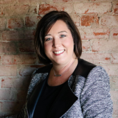 Headshot of Kristin Winkle Beck, She is standing in front of a brick wall, wearing a black and grey blazer