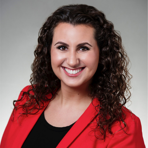 Headshot of Natalie Hales, she has curly brown hair and a big smile, wearing a red blazer