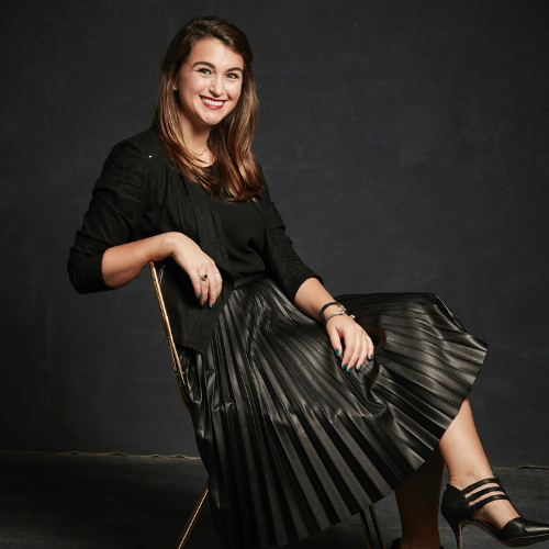 Headshot of Taylor Anne Adams, she is sitting in a chair with her legs crossed and arm draped across the back of the chair. She is wearing a shiny skirt and big smile
