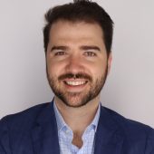 Man with dark hair and beard in blue shirt with blazer