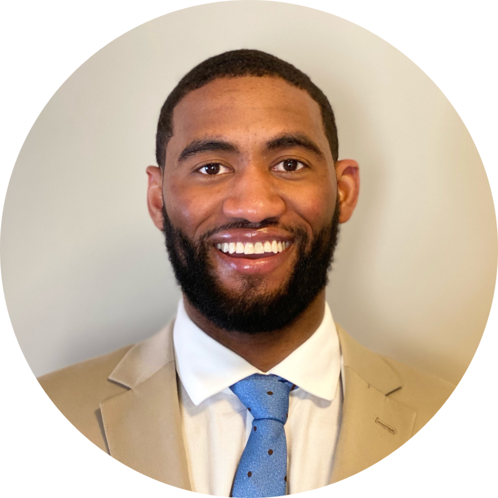 Headshot photo of man with beard smiling and wearing a white shirt, blue tie, and khaki jacket