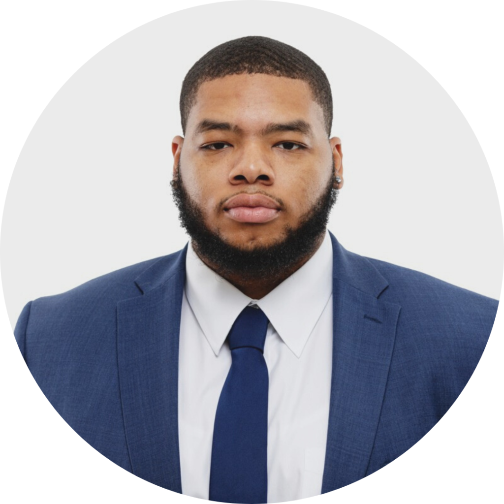 Headshot photo of man with dark board wearing a navy suit, white shirt, and blue tie