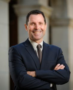 Tony stands with arms crossed wearing a dark suit in front of a series of arches on campus