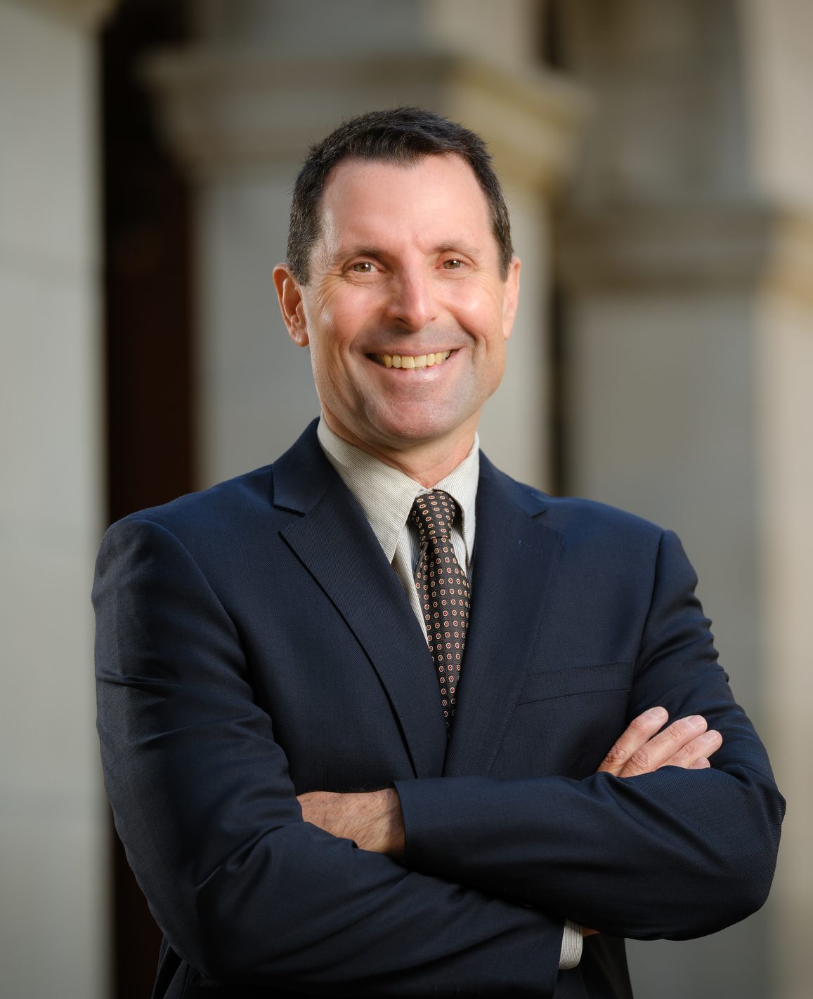 Tony stands with arms crossed wearing a dark suit in front of a series of arches on campus