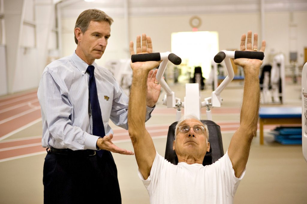 Professor Jim Ross watches over a patient as the patient raises a workout bar over his head.