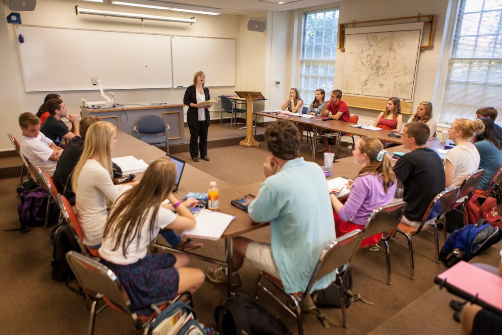 Kathy Smith stands in the center of a u-shape of desks as students look at her.