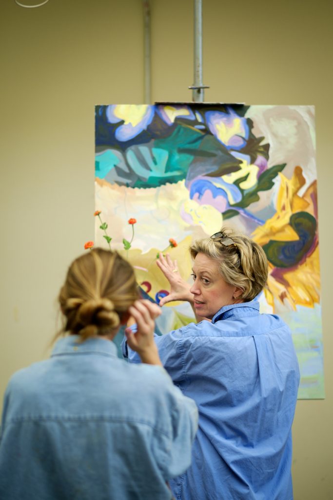 Page Lauglin looks over her shoulder to student whose back is to the camera as they discuss a painting.