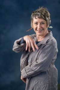 Lynn Book dressed in a grey shirt waves in front of a dark grey background