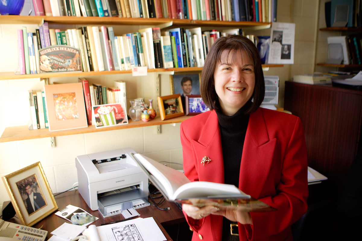 Claudia Kairoff wears a red blazer and holds a book in her office