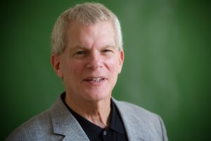 Michael Hyde headshot. He wears a grey suit in front of a blackboard.