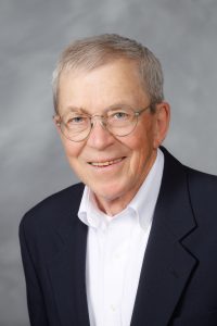 John Wood sits for a headshot in a black suit on a grey background.