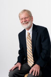 Professor Bill Connor sits for a headshot. He is wearing a black suit and yellow and black tie.