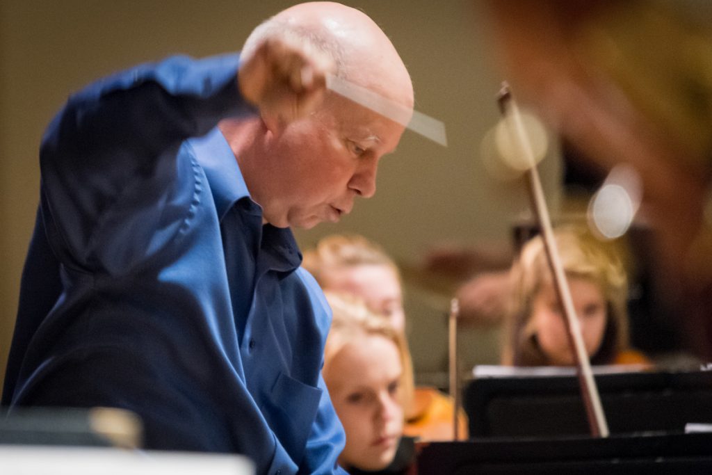 David Hagy looks at his sheet music with a raised baton. He is wearing a blue collared shirt.