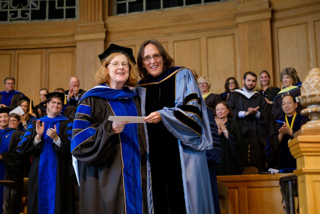 Classics professor Mary Pendergraft, wearing regalia, receives the Award for Excellence in Advising from Christy Buchanan