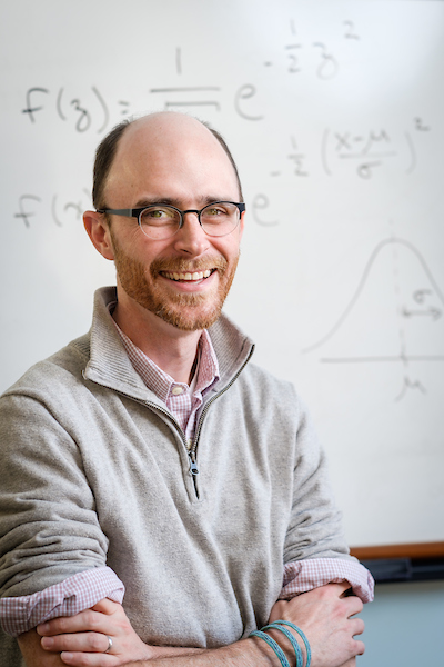 Rob wears a tan sweater and glasses as he sits arms crossed in front of a white board with an equation on it.