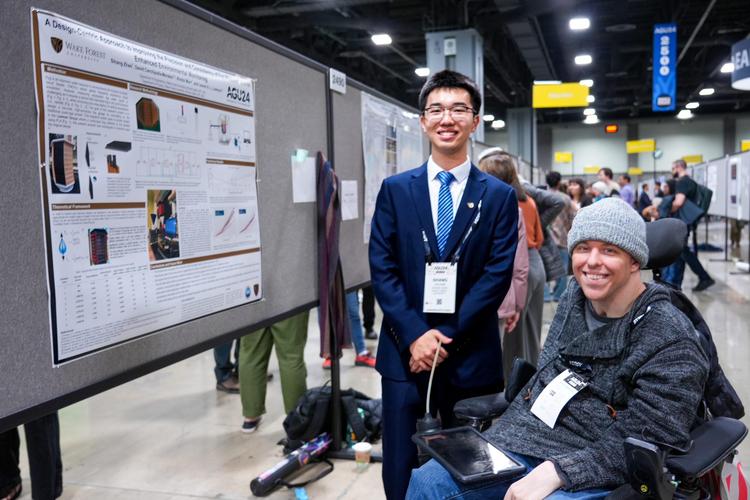 Cosmo Zhao stands next to his poster presenting his research in a crowded room of other poster presentations at the conference. 