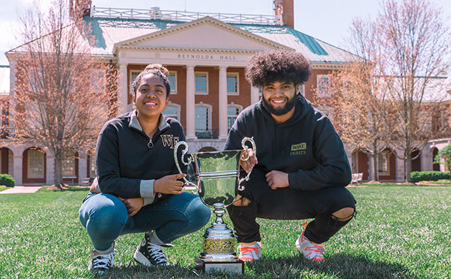 Two students posing with debate trophy in front of Reynolda Hall