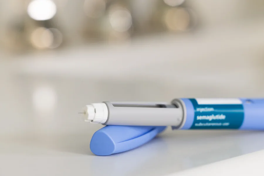 GLP-1 semaglutide injection pen, a medication used to treat Type 2 diabetes and aid in weight loss, on a white surface. The prescription medicine is used to treat Type 2 diabetes as It helps regulate blood sugar levels by stimulating insulin secretion and decreasing glucagon levels. In addition, it is also used off-label for weight loss in individuals with obesity, as it can help reduce appetite and increase feelings of fullness. The pen is positioned horizontally, with its cap removed, on a bathroom countertop with a white surface. The medical device is in the foreground and the background is out of focus because of a shallow depth of field.