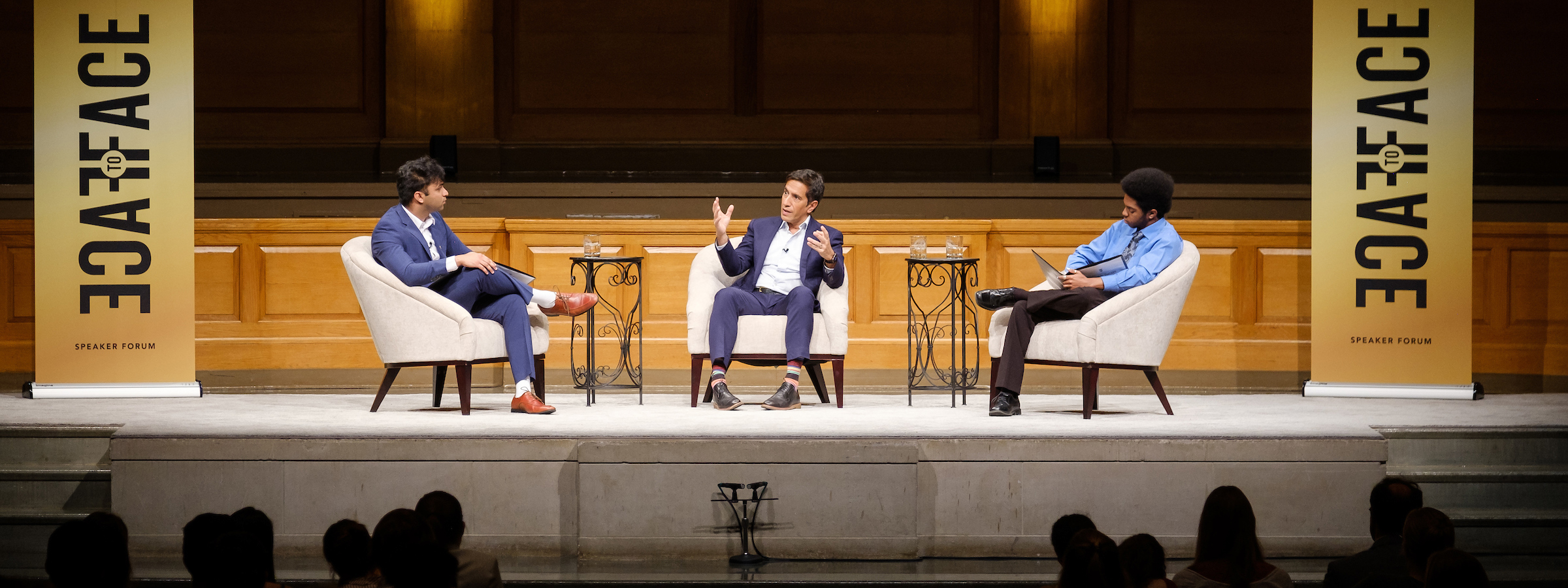 Dr. Sanja Gupta sits with two other men on the stage of Wait Chapel in front of a seat crowd