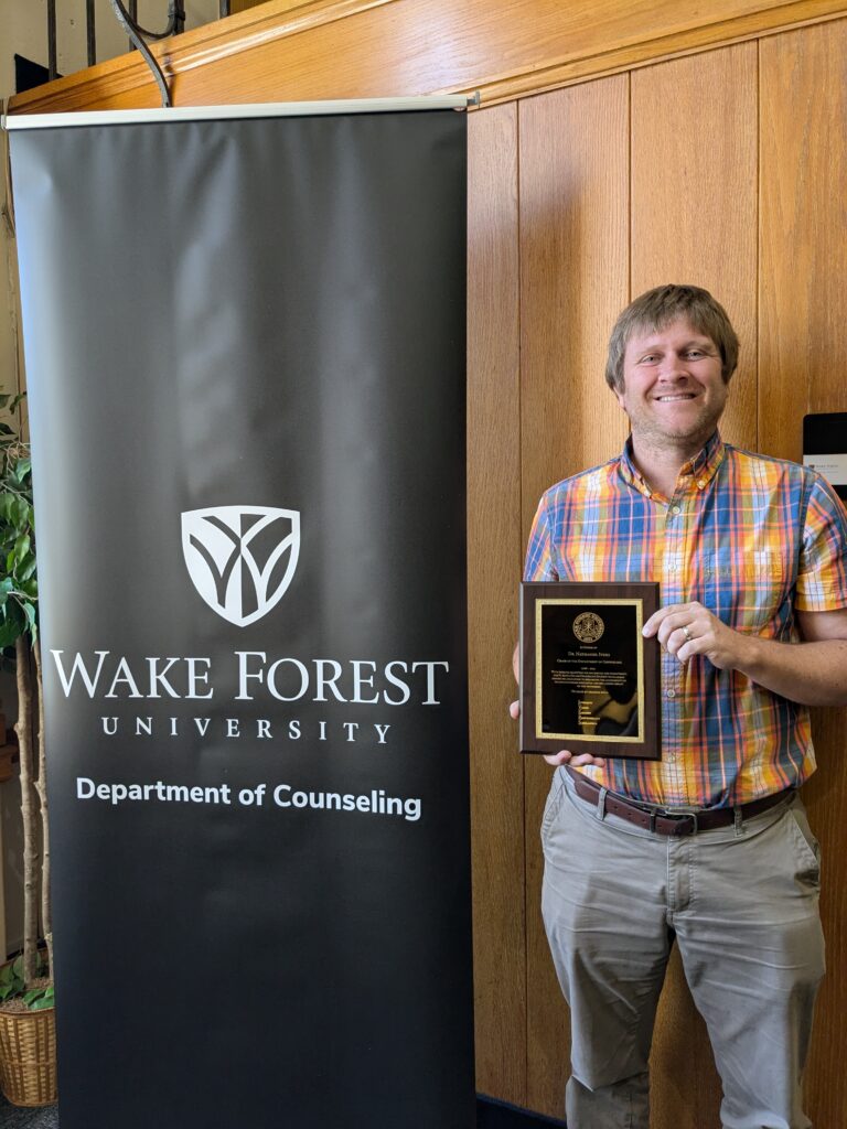 Dr. Nate Ivers holding plaque commemorating new Pi Alpha scholarship with Department of Counseling banner.