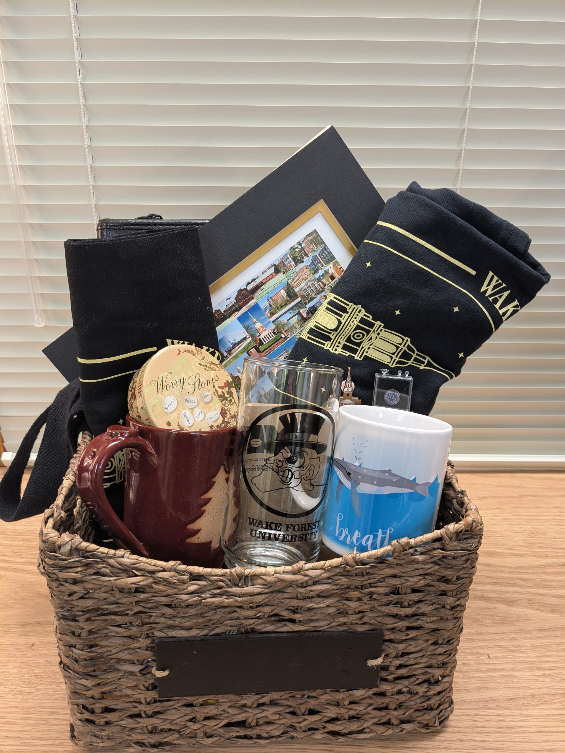 Pi Alpha Raffle Drawing. Photo of Basket #2 'Pi Alpha and Self-care'. Includes a black leather CSI portfolio, a vintage glass; a matted photo of various sites on campus; a WFU Wait Chapel pin, a CSI pin, a WFU journal and pen set; a WFU cloth grocery bag and WFU t shirt; a pottery coffee mug, a 'Breathe' coffee mug and 6 stress relief toys. Value at $100+