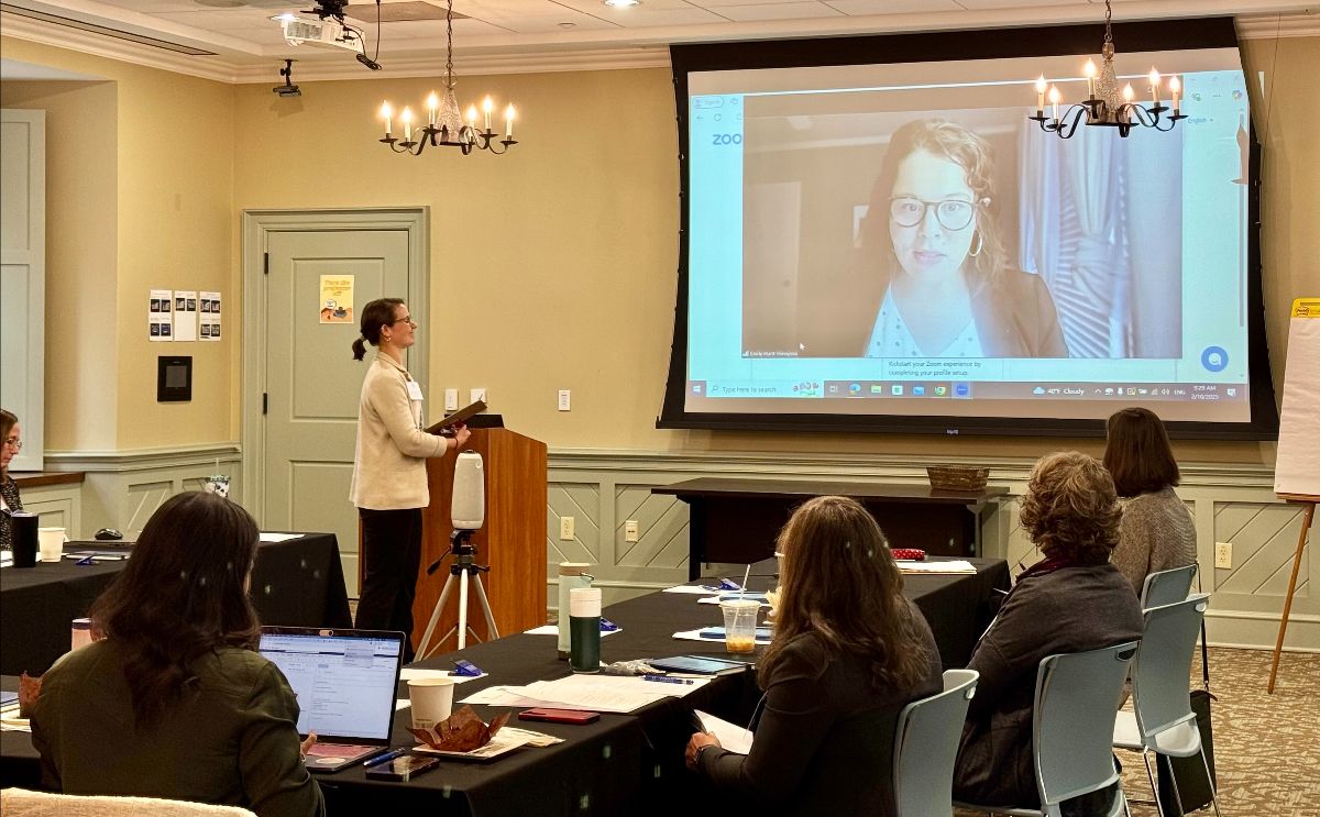 Liz Whiting stands at a podium while Emily Hunt-Hinojosa speaks to the conference via Zoom.