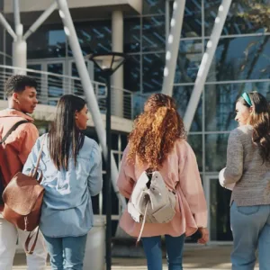 Education campus, students and walking at college to class with talking, conversation or exam chat. Back, friends and diversity outdoor at university with backpack for learning, scholarship and study.