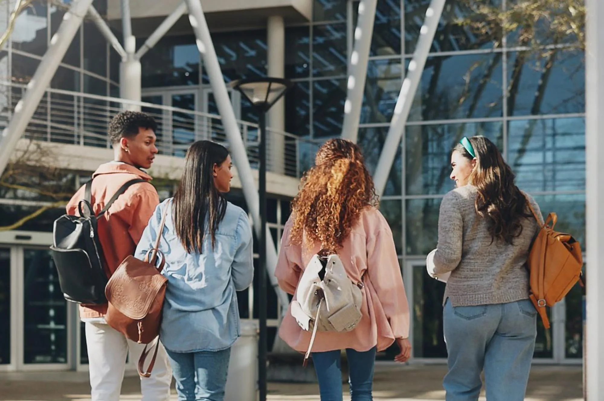 Education campus, students and walking at college to class with talking, conversation or exam chat. Back, friends and diversity outdoor at university with backpack for learning, scholarship and study.