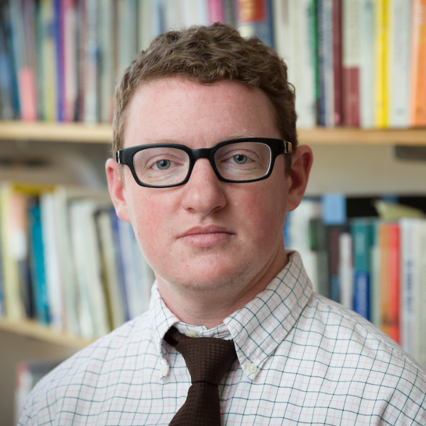 Person in front of bookshelf wearing black glasses and a button-up shirt and tie