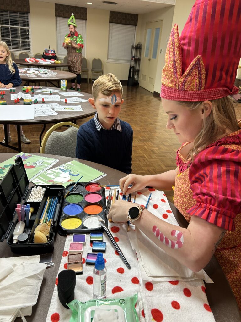 A volunteer dressed as an elf in colorful holiday attire paints a child's face at the WAKEAtlanta Lovefeast gathering.