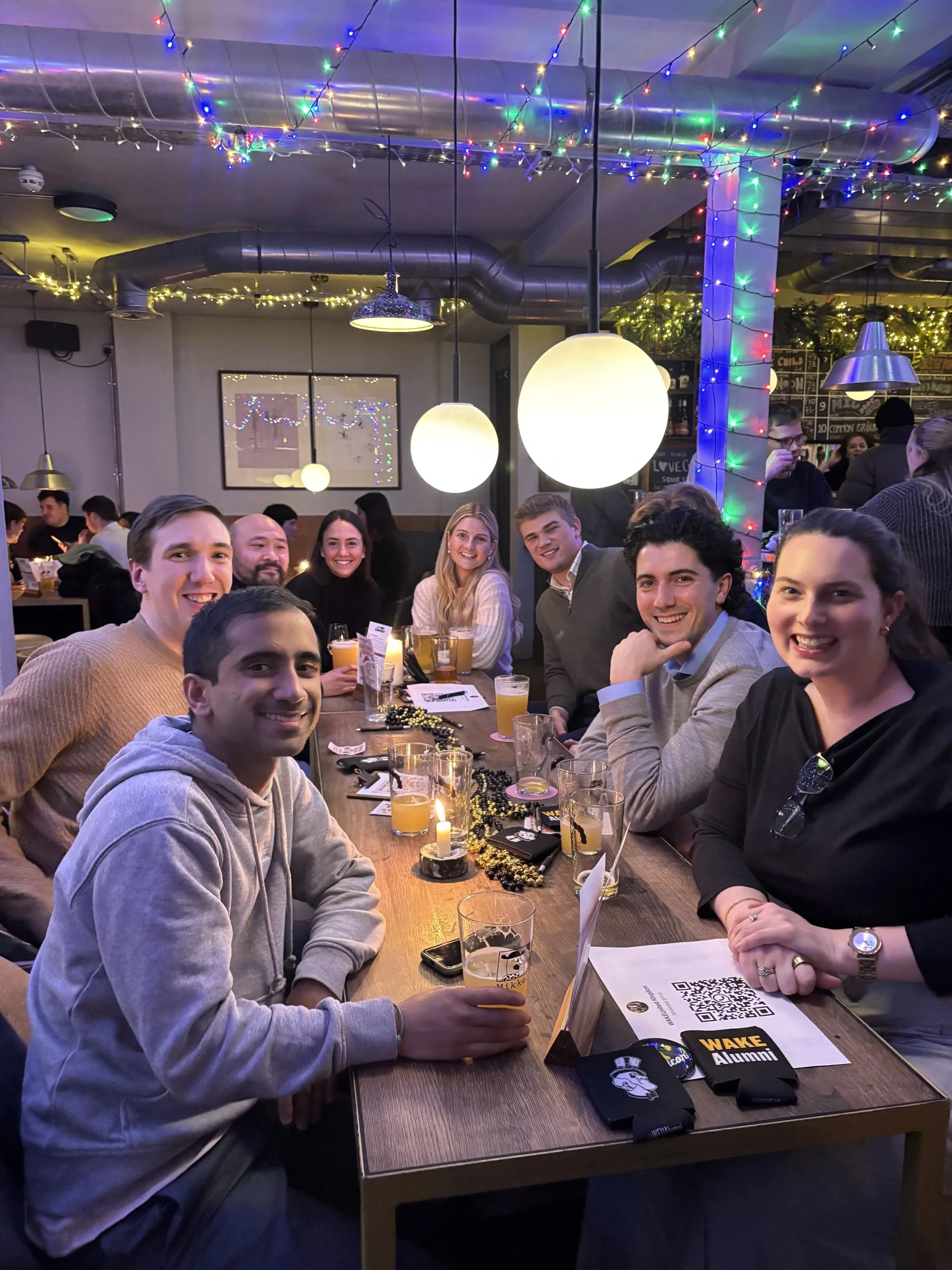 Wake Forest alumni sit at a long bar table facing the camera and smiling. The table is covered with pints of beer, black and gold beads, and Wake Forest koozies. Multicolored string lights are hung up along the ceiling above the group and wrapped around a pillar behind them.