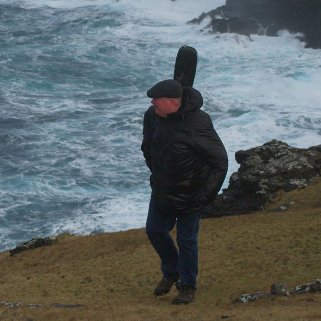 A man with a guitar on his back walks near the shore.