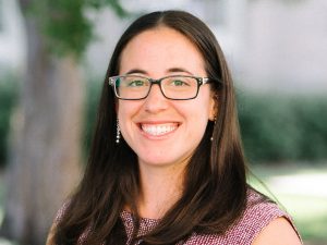 Portrait of Gia Branciforte. Gia is a white woman with straight brown hair. She is wearing glasses, silver ball earrings, and a patterned dress and is smiling at the camera.