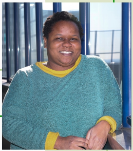 A photo of a Black plus-sized woman with short sponge-like natural hair, wearing a mid-toned sweater with bright accents, smiling warmly. The background is a soft, neutral scenery with good lighting. The style is realistic and casual.