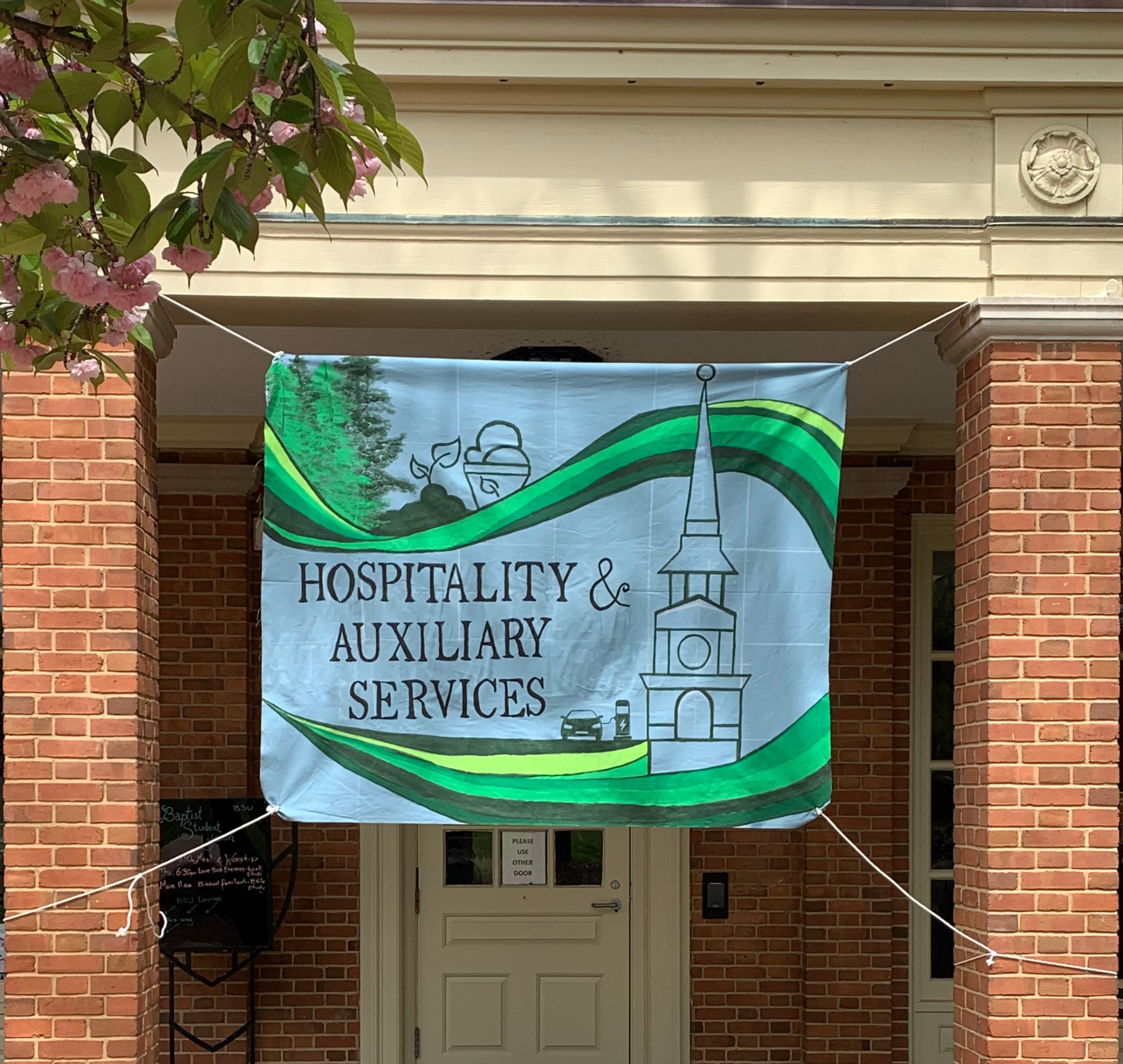 Hand painted canvas banner highlighting sustainability efforts by Hospitality & Auxiliary Services hanging on campus. 