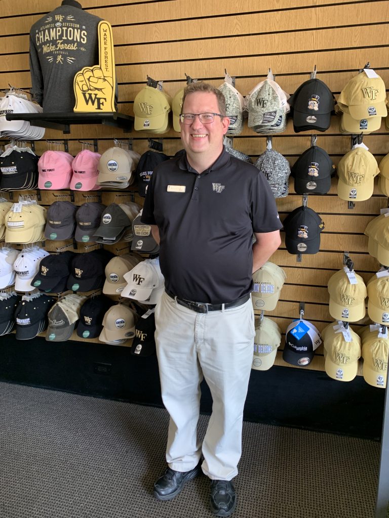 Photo of Deacon Shop Manager, James Hamilton, standing in front of the hat display in the Deacon Shop on campus.
