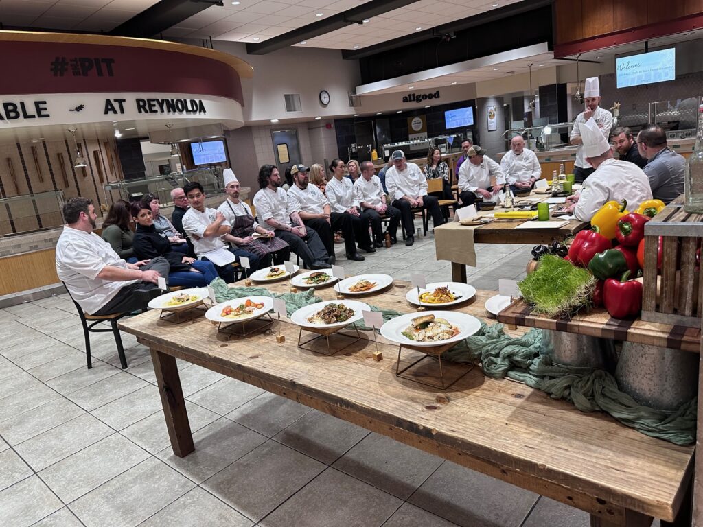 A group of chefs gathered for a culinary presentation or judging event, with a variety of prepared dishes displayed on a table in the foreground.