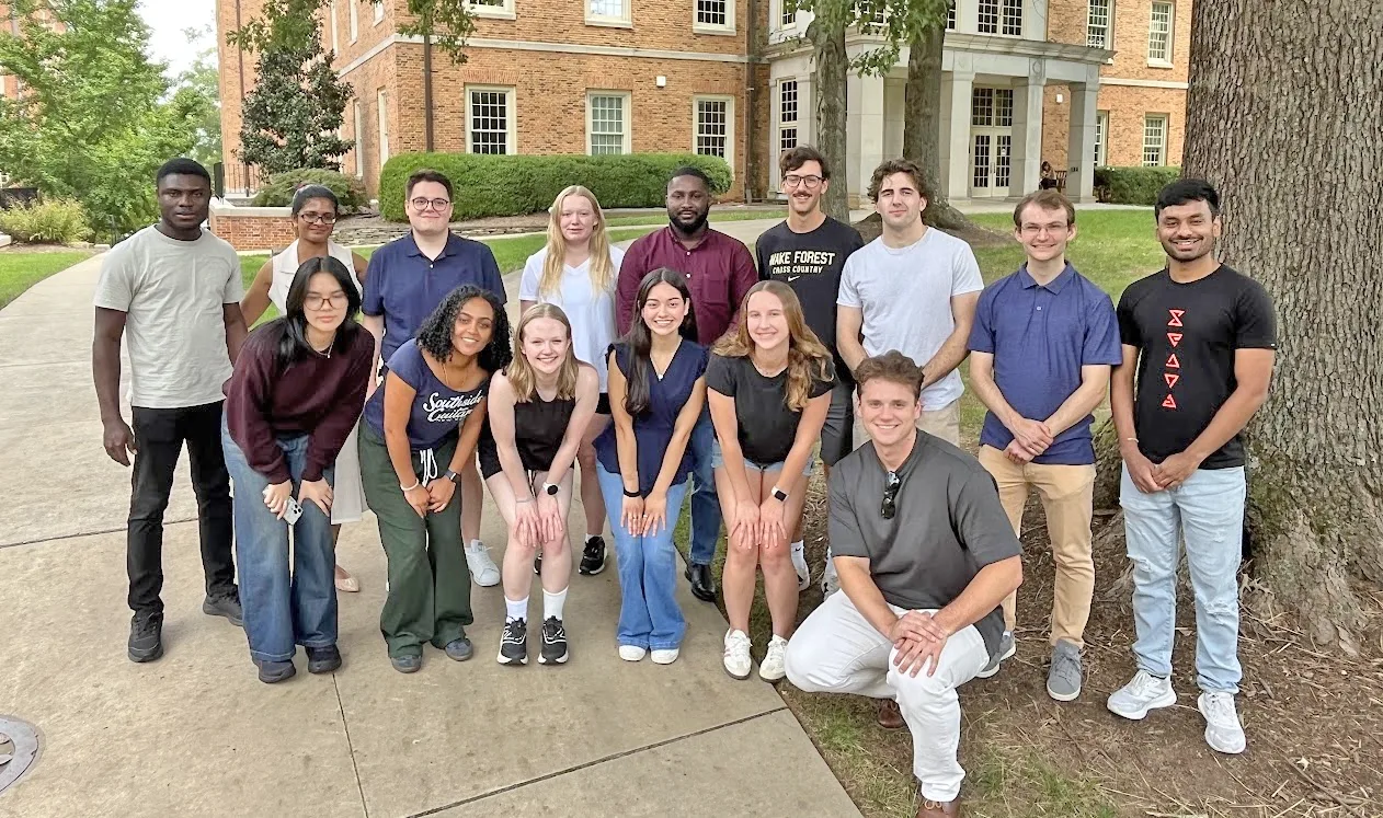 Group of students under a tree, campus buildings in background