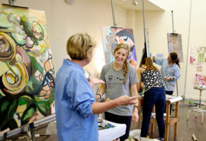 Studio painting class led by Page Laughlin, art department chair and professor of painting, September 2014. A student smiles at Laughlin gesturing in front of paintings on the wall and easels.