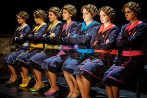 A row of women in bobbed curvy 1940s hairdos in colorful uniforms sit primly in chairs in a dress rehearsal for "The Adding Machine," directed by Professor Brook Davis, on the Tedford Stage, February 2018.