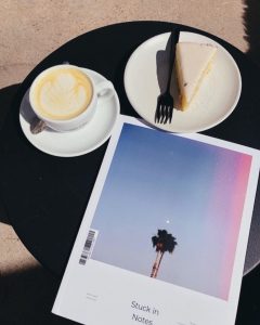 First issue of Stuck in Notes magazine with cover of solitary California tree and moon on a table with a cup of latte and a plate with cheesecake and fork shot from above on a black placemat