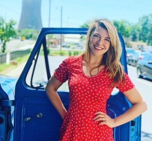 Devin Kidner (08) in a red print dress in front of an open passenger side door along a street.