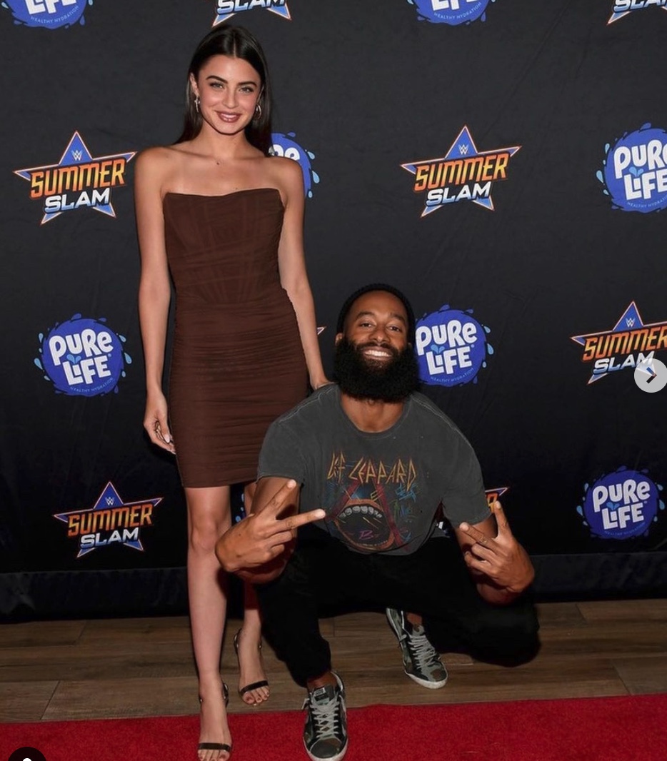 Matt James ('14) with his girlfriend Rachael Kirkconnell in a photo shoot for an event. She's in a strapless brown dress and he's giving peace signs as he squats. He still has his bushy long beard that he recently shaved off.