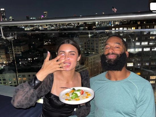 Matt James ('14) sits on an outdoor sofa at night with girlfriend Rachael Kirkconnell as she holds a plate of food and licks her fingers.
