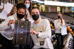 Two women's basketball players hold up a poster that reads "WE ARE IN!" after being selected to the NCAA Tournament.