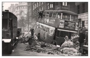 A bus that reads "Picture Post, 77, Kings Cross" sits in rubble. A rope is thrown up to second floor passengers.