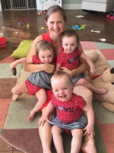 Jill sits on the floo with two triplets in her arms and one in between her legs, all smiling up at the camera