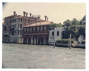 Old photo of what became the Casa Artom building. The building is dark brown with lots of white poles in front. The building is on the Grand Canal.